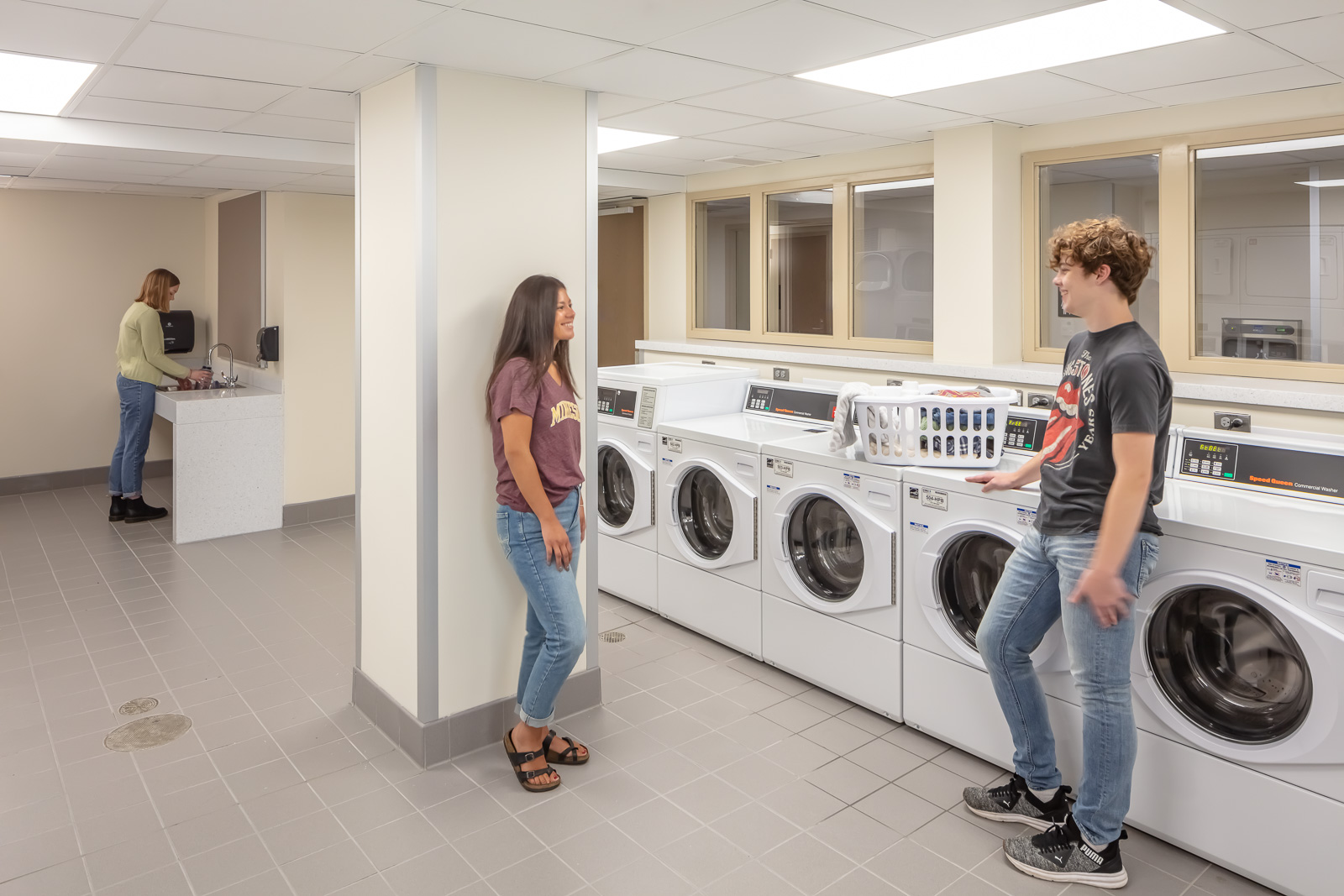 Two students stand beside each other and talk in an open, spacious laundry room alongside a row of washing machines. In the far left, a woman washes her hands at a sink.