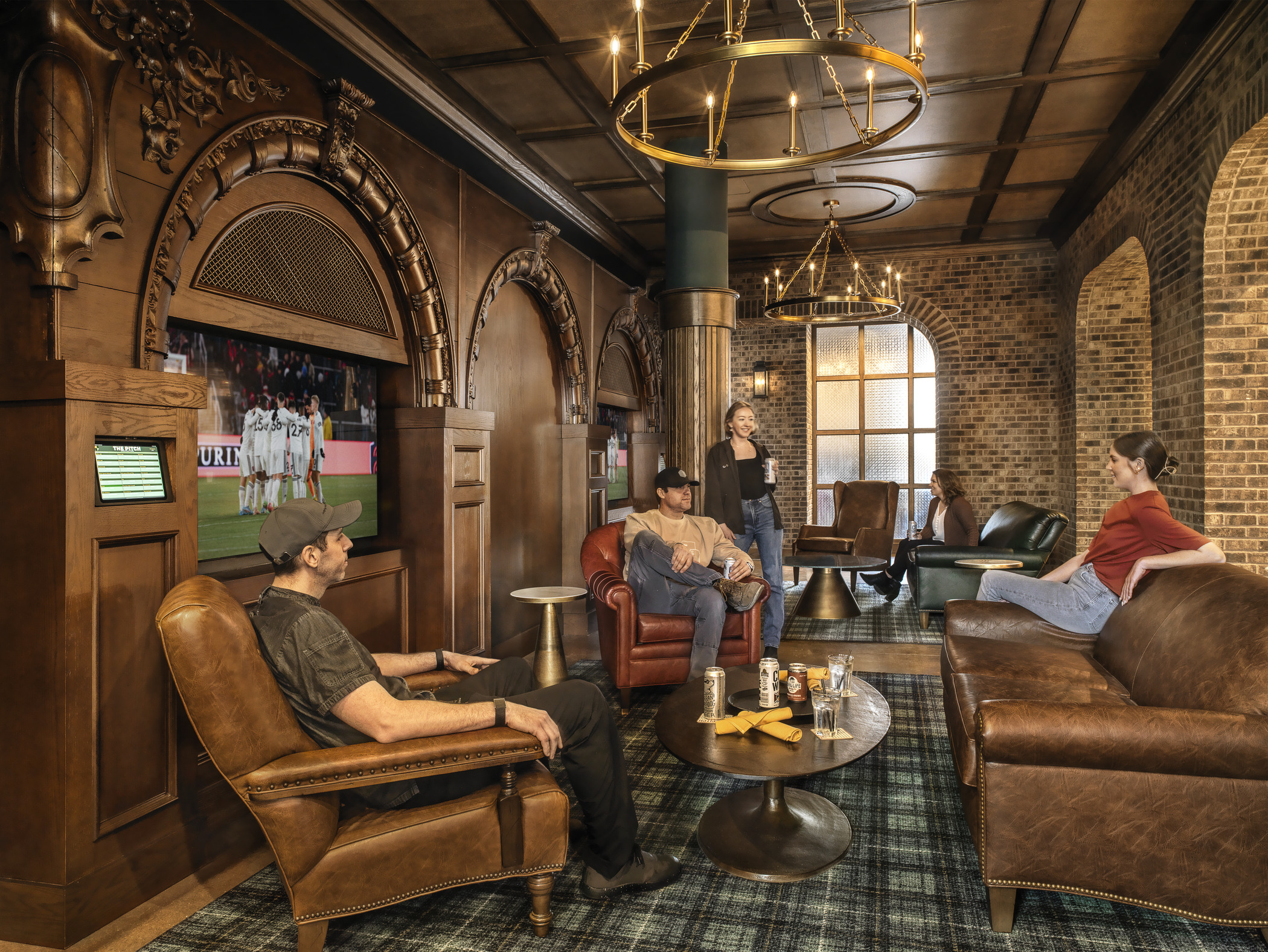 A group of 5 people relax in a lush lounge space, with soccer matches playing on TVs in recessed parts of the space's wall
