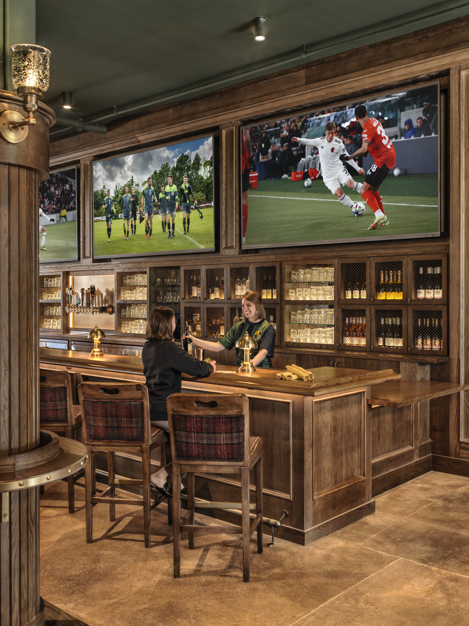 A woman in a soccer-themed outfit serves a beer to a customer inside an ornate wooden bar, as soccer matches play on TVs above the bar