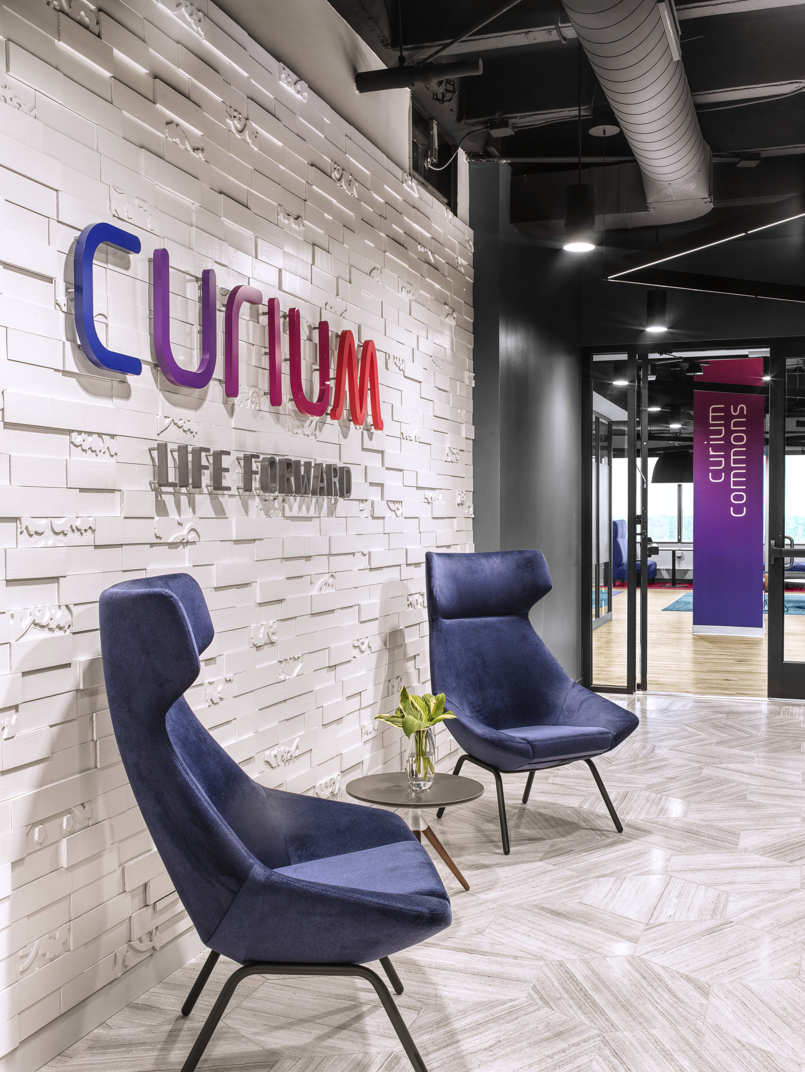 Two deep-set fabric chairs sit in a bright, clean lobby area. On the wall behind them, text reads "Curium. Life Forward." A doorway to the left leads to an open working space, with the text "Curium Commons" printed on a painted support beam matched to the Curium colors.