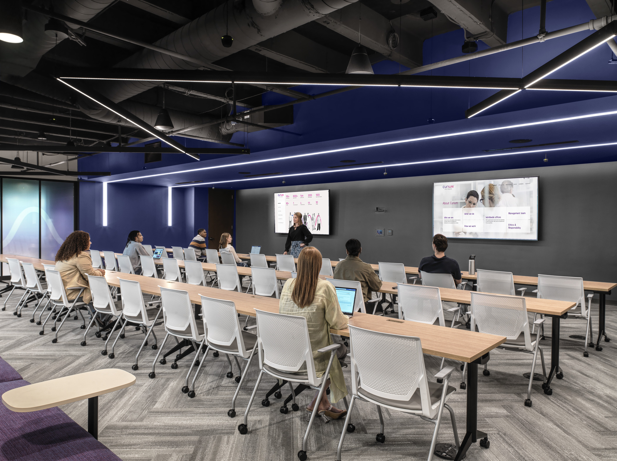 A variety of young people sit in a spacious multipurpose room, with a woman at the front of the room standing in front of two screens branded to Curium graphics. Above, sleek and modern light strips create geometric patterns in the ceiling.