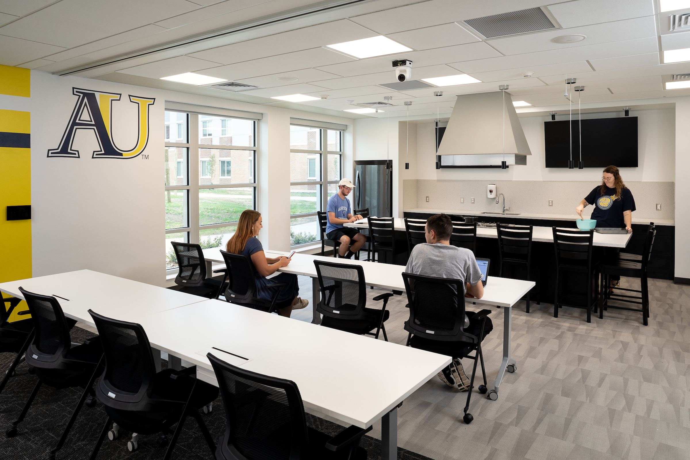 A variety of students sit, talk, study, and cook food inside a residential hall multipurpose room.