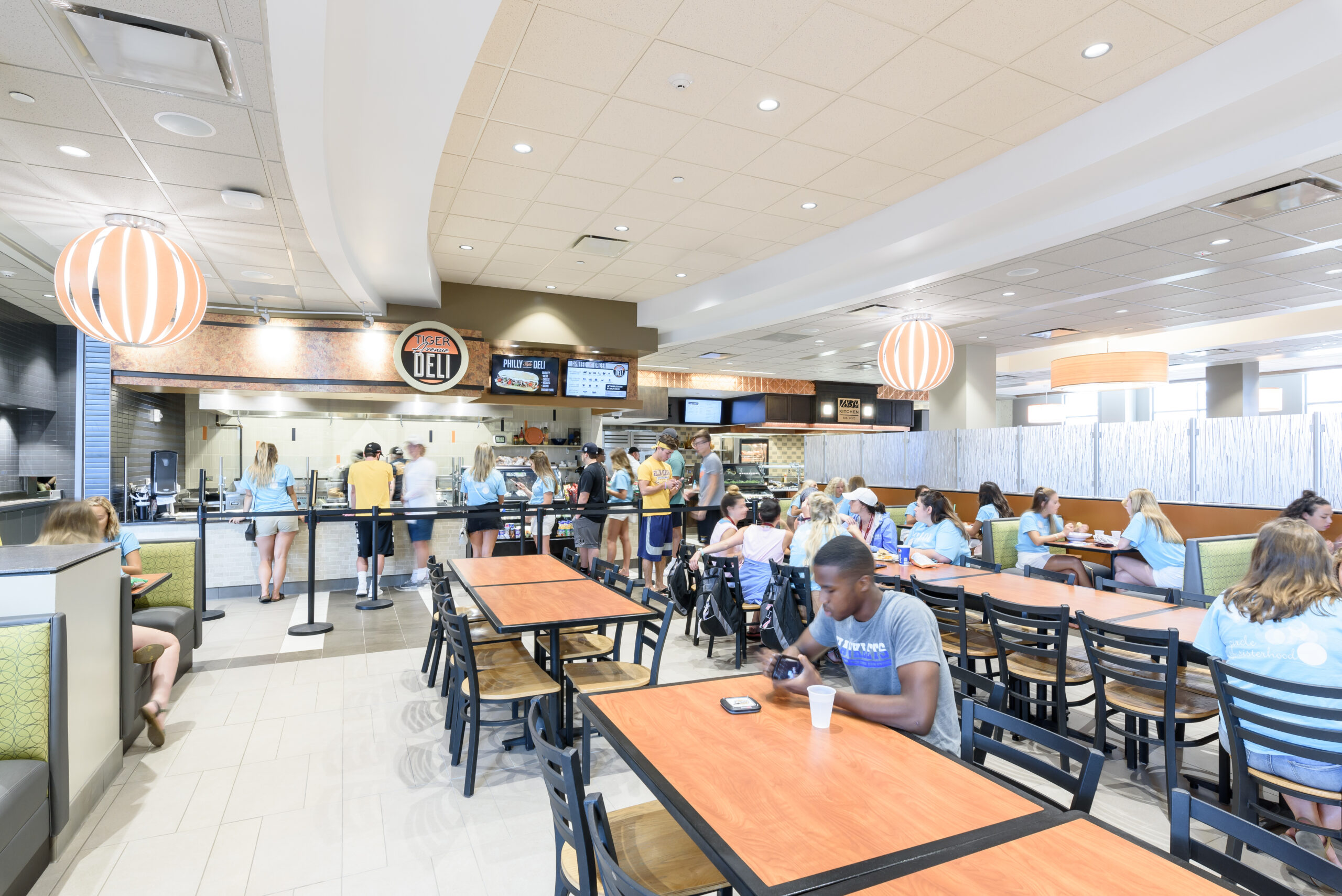 A large number of college students occupy a dining hall, with a station named "Tiger Avenue Deli" serving a large line of people