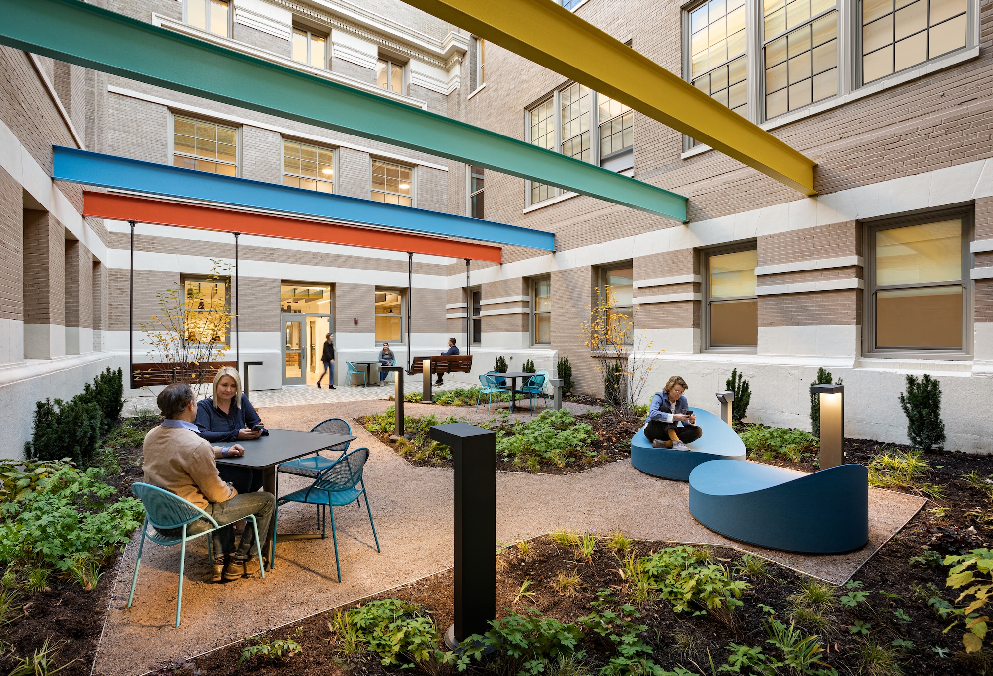a photograph of an outdoor patio with multiple people at tables. overhead are colorful railway beams