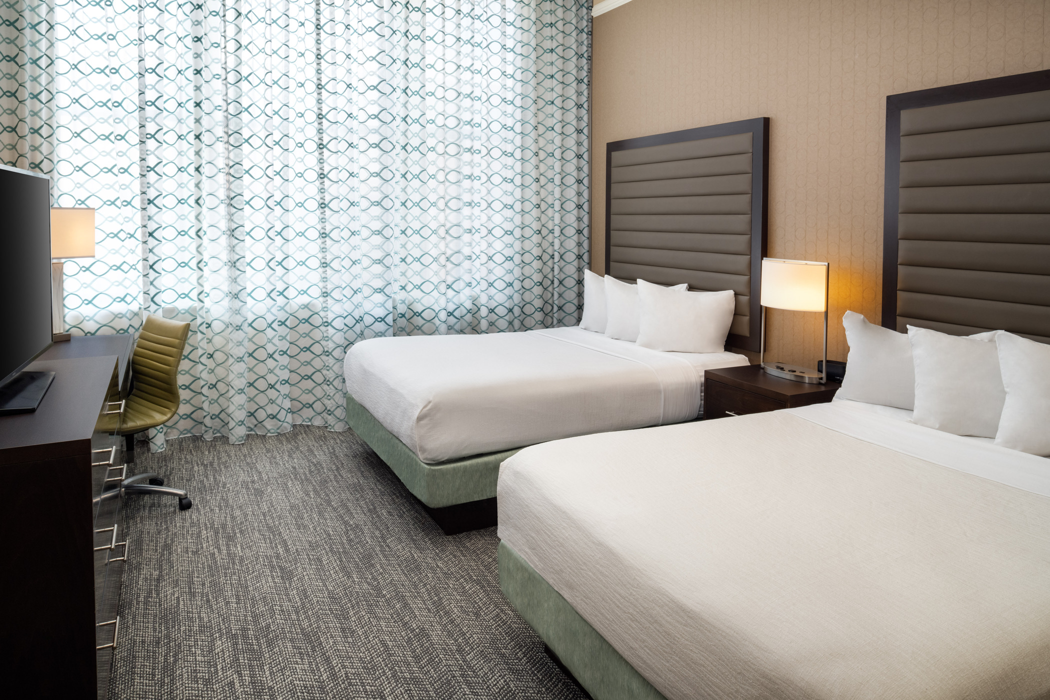A hotel room with two beds neatly made with white sheets and brown tall headboard in front of a window.