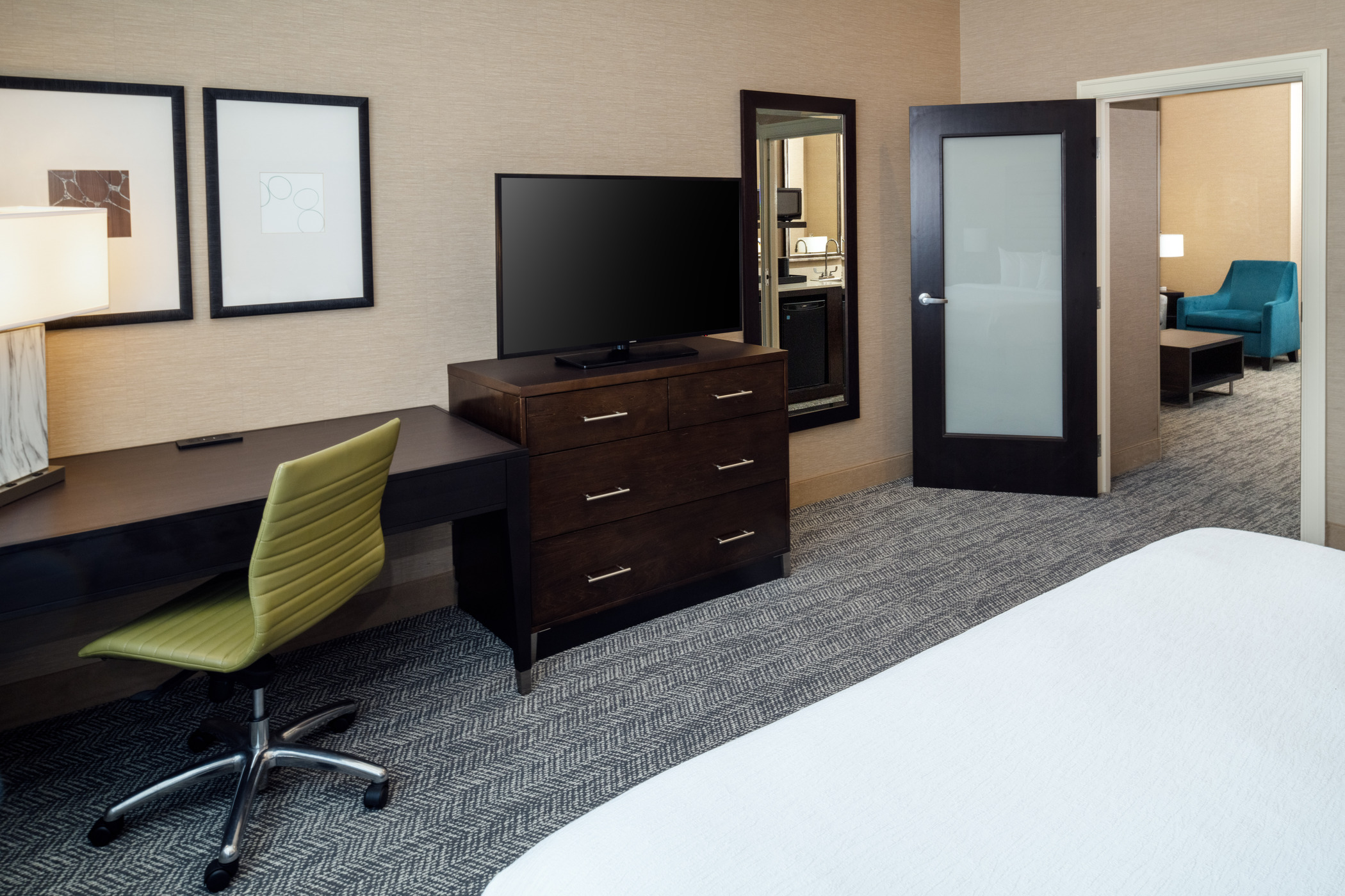 A view from a bed facing a dresser and a TV on top, to the left is an attached desk and a chair.