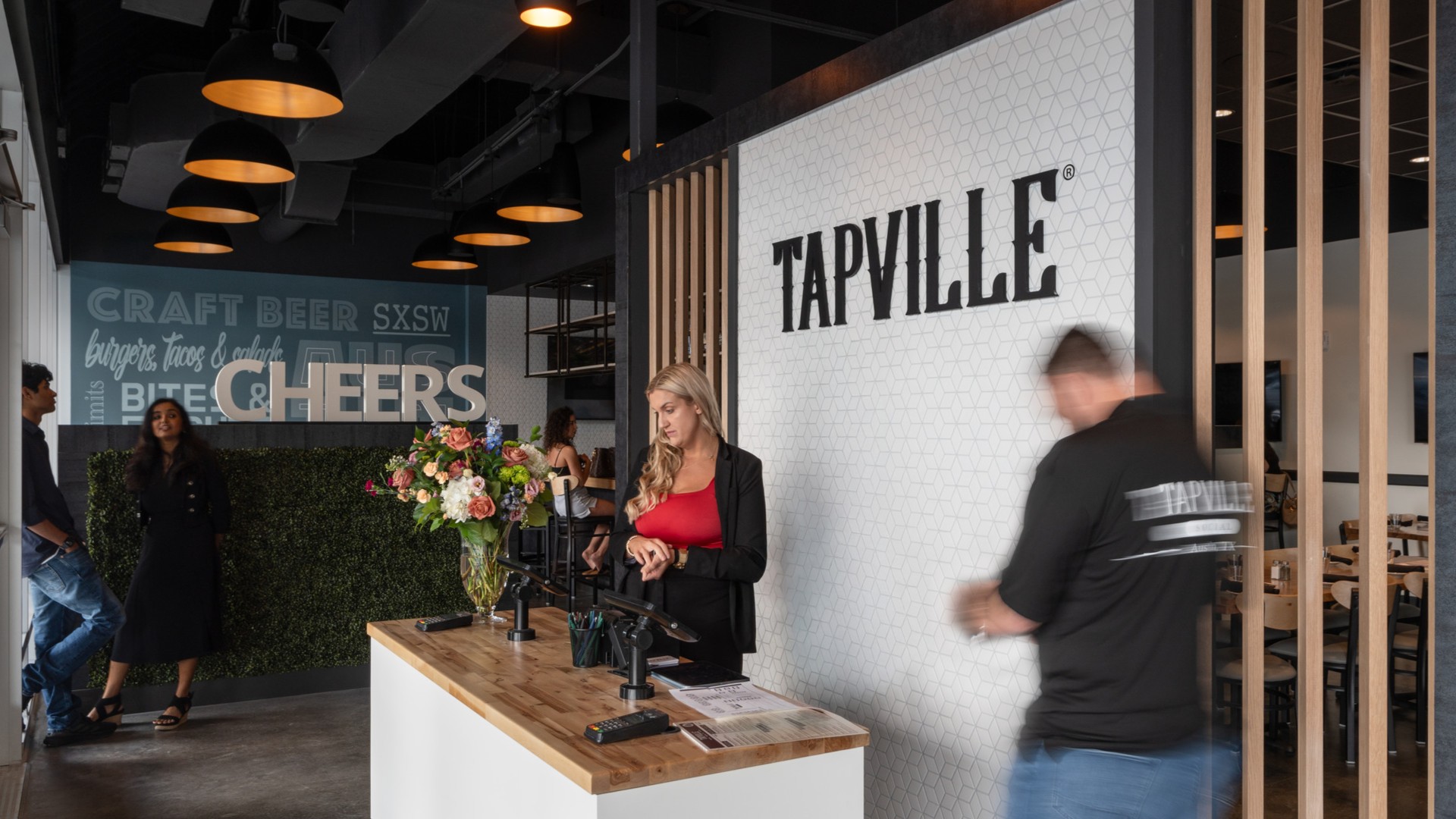 A server walks towards a hostess in front of a standee wall which displays the Tapville logo.