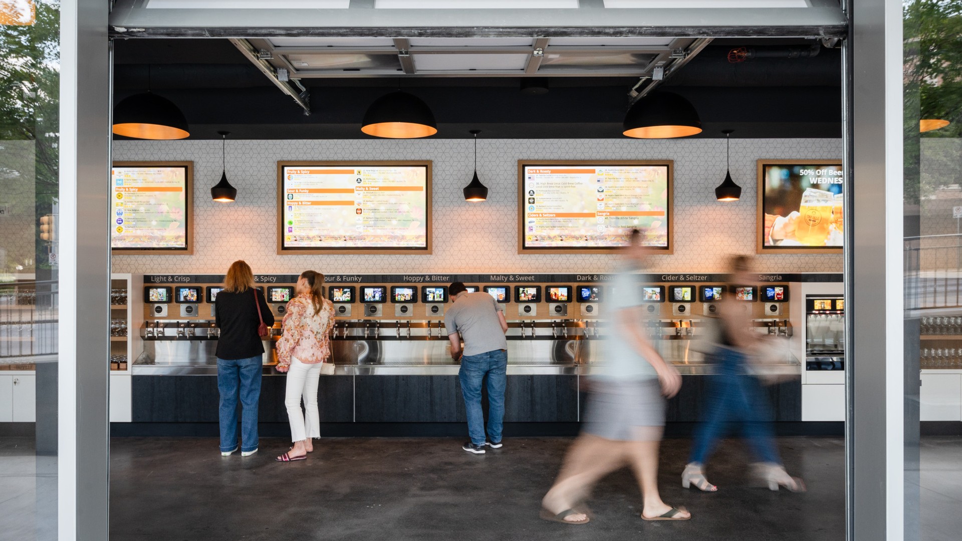 A group of people walk and are seen pouring beer from a wall of self-serve beer taps, photographed through an indoor-outdoor garage door