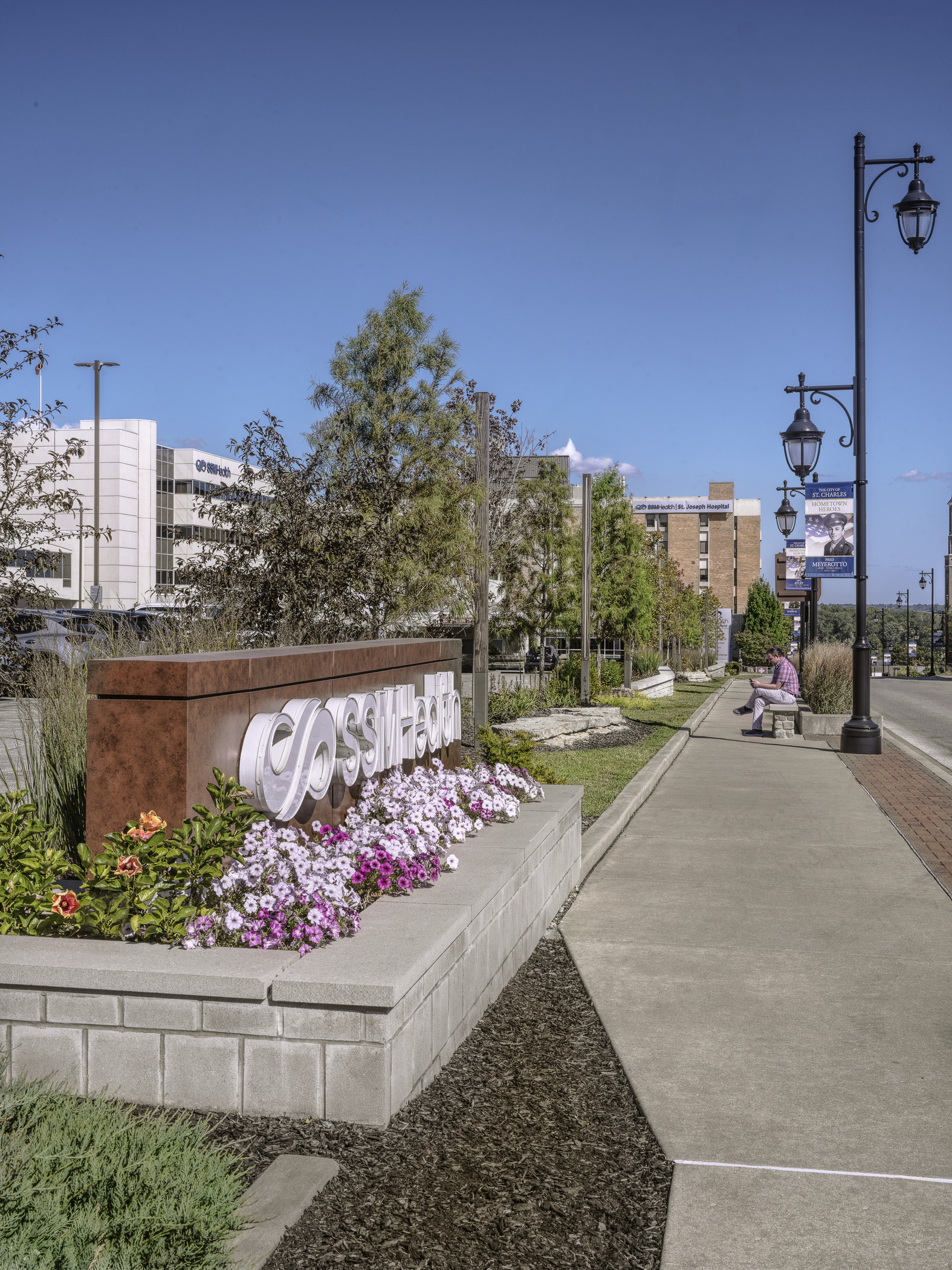 A hospital campus and the surrounding streetscape is photographed from the public sidewalk at daytime. The SSM Health logo is visible via a marquee sign in the foreground of the picture.