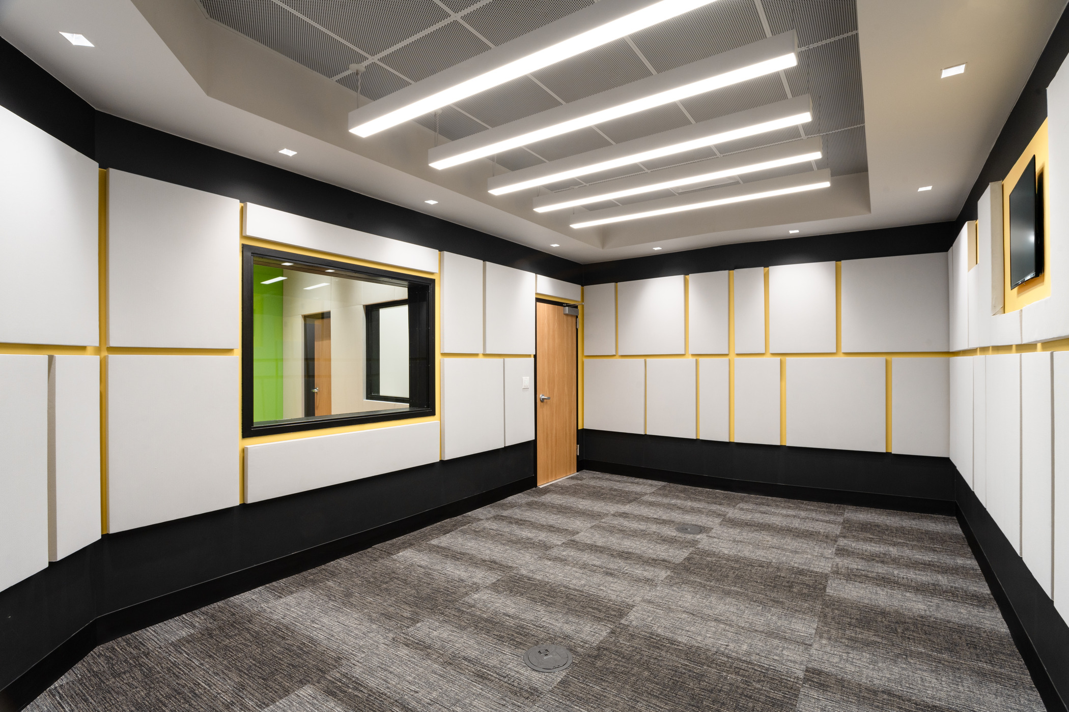 A modern large room with white soundproof boards on the walls with yellow paint seen in between boards. A row of overhead lights on the ceiling and gray flooring. A connecting wall with a door and a window to see through the next door room.