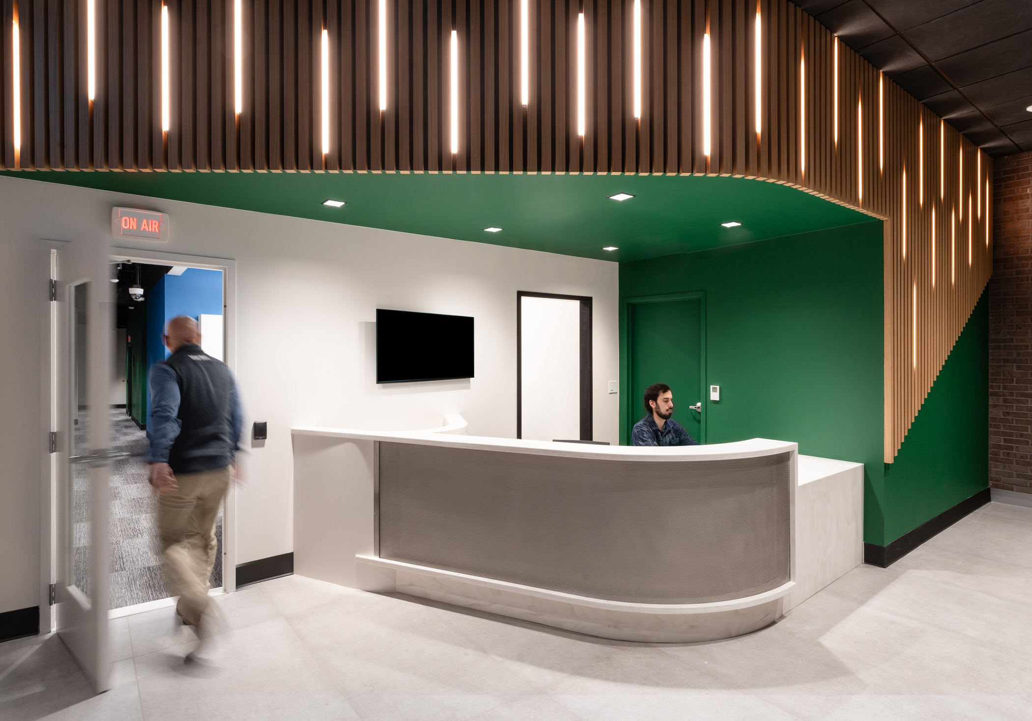 Modern reception area with a curved front desk, a receptionist seated behind it, and a person walking through an open door on the left. The space features a green accent wall, wood slat lighting details overhead, and a mounted blank monitor on the wall.
