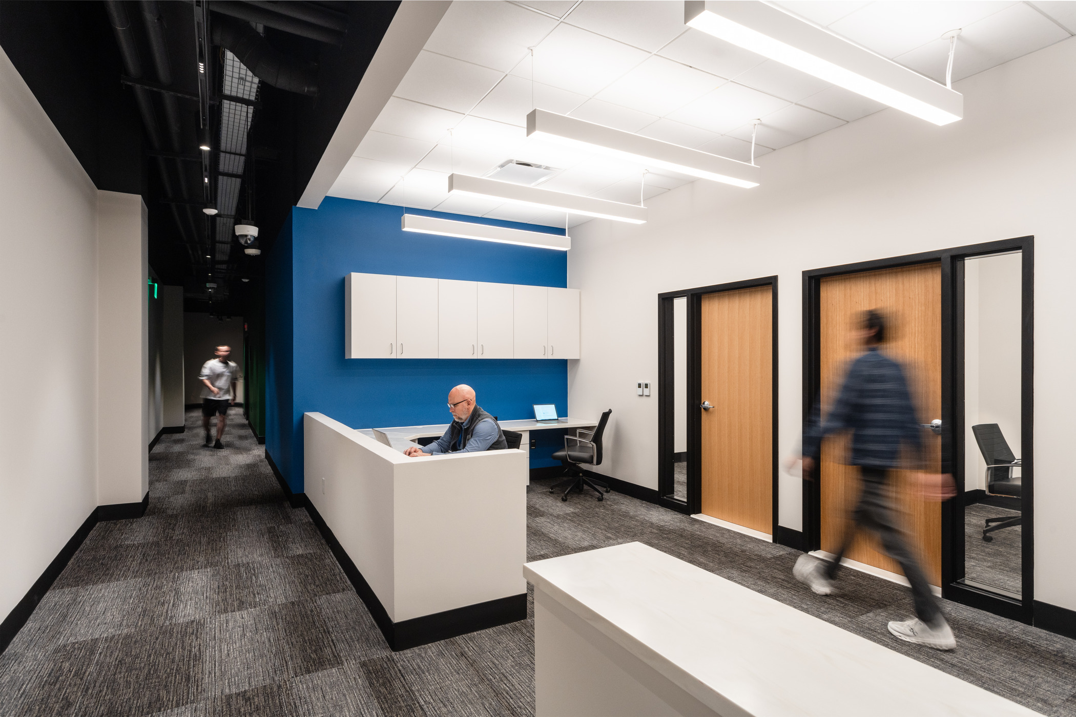 Office hallway with a small reception desk where a person is seated working on a laptop. Two people walk through the corridor, one approaching from the distance and one passing by on the right. The space features wood-finish office doors, a blue accent wall with white cabinets, and modern linear ceiling lights.
