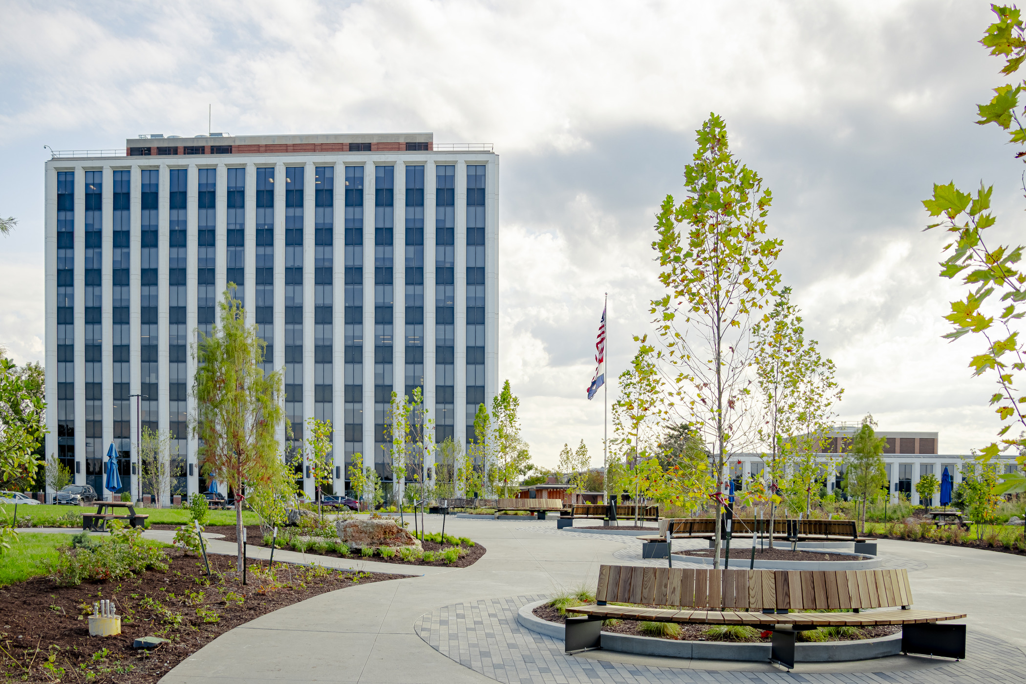 View of 1385 Plaza featuring circular wooden benches around trees, lush greenery, and a tall corporate office building in the background, showcasing modern commercial landscape design.