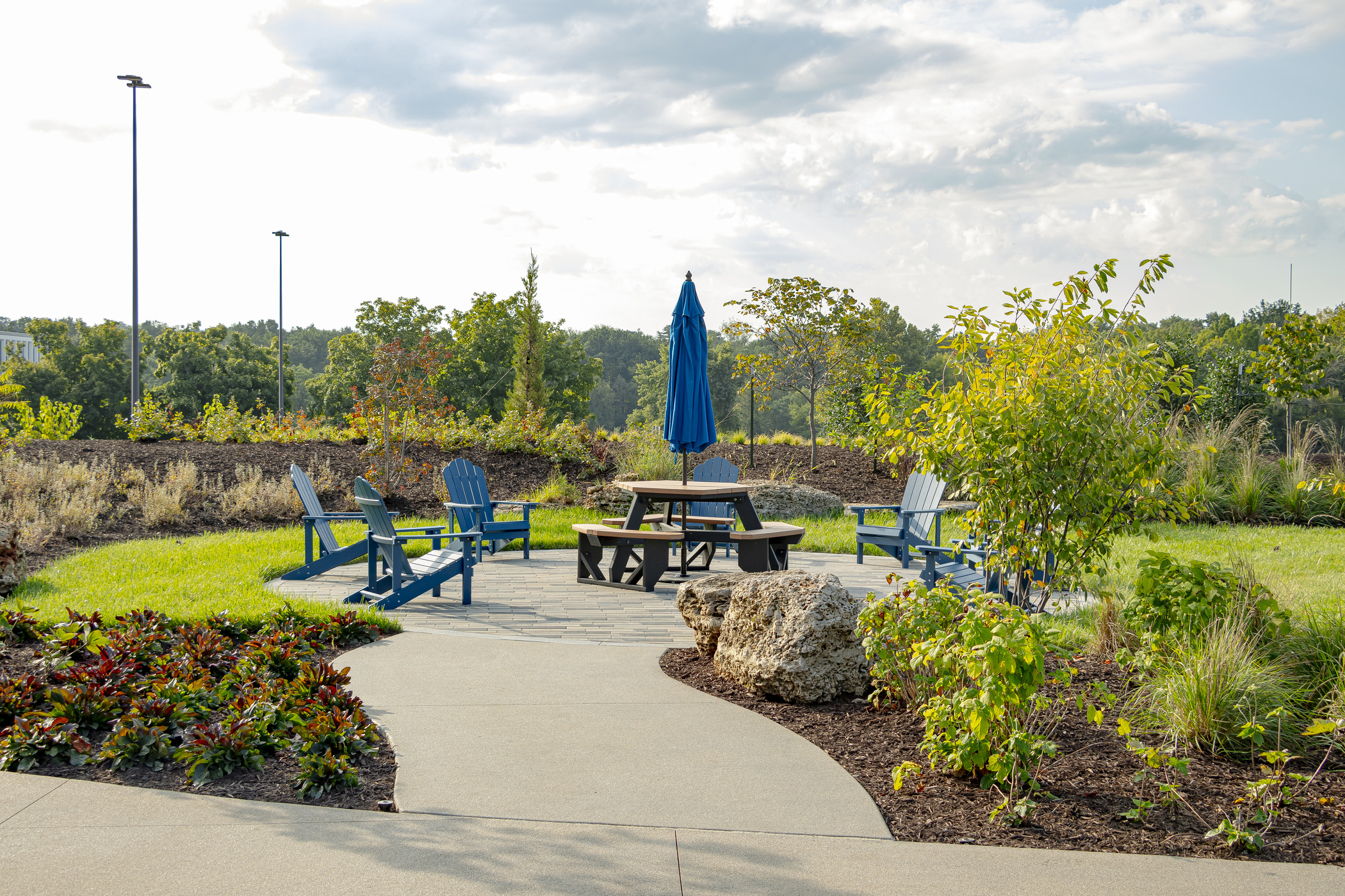 Cozy outdoor lounge area with blue Adirondack chairs, umbrella, and picnic table surrounded by native plants and stone accents at Maritz Campus 1385 Plaza, promoting connection to nature.