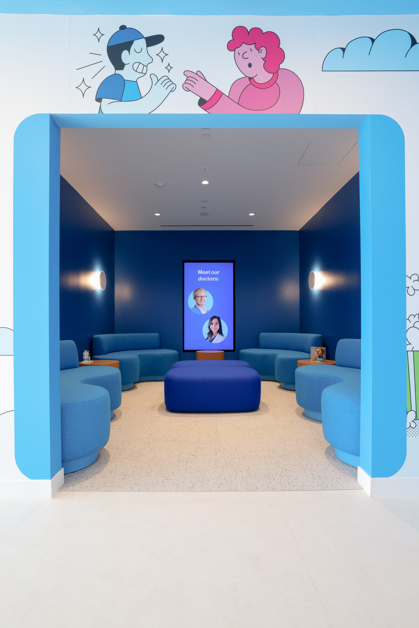 Dental office lounge with deep blue walls and matching seating arranged around a central ottoman. A digital screen displays ‘Meet our doctors,’ and playful cartoon graphics decorate the entrance, creating a friendly and modern waiting area.