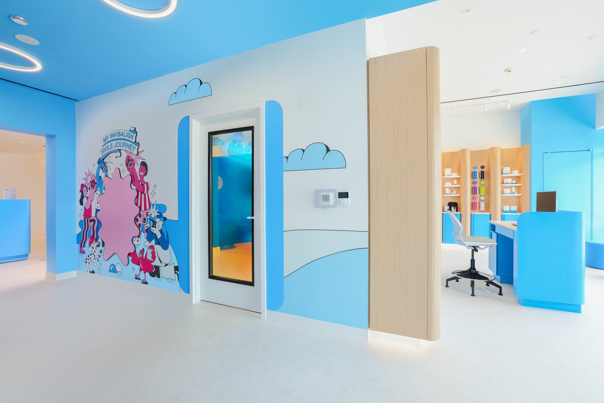 Dental office interior with bright blue and white walls featuring playful cartoon graphics and the phrase ‘My Invisalign Smile Journey.’ A glass door leads to another room, and a modern reception area with wood accents and blue counters is visible.