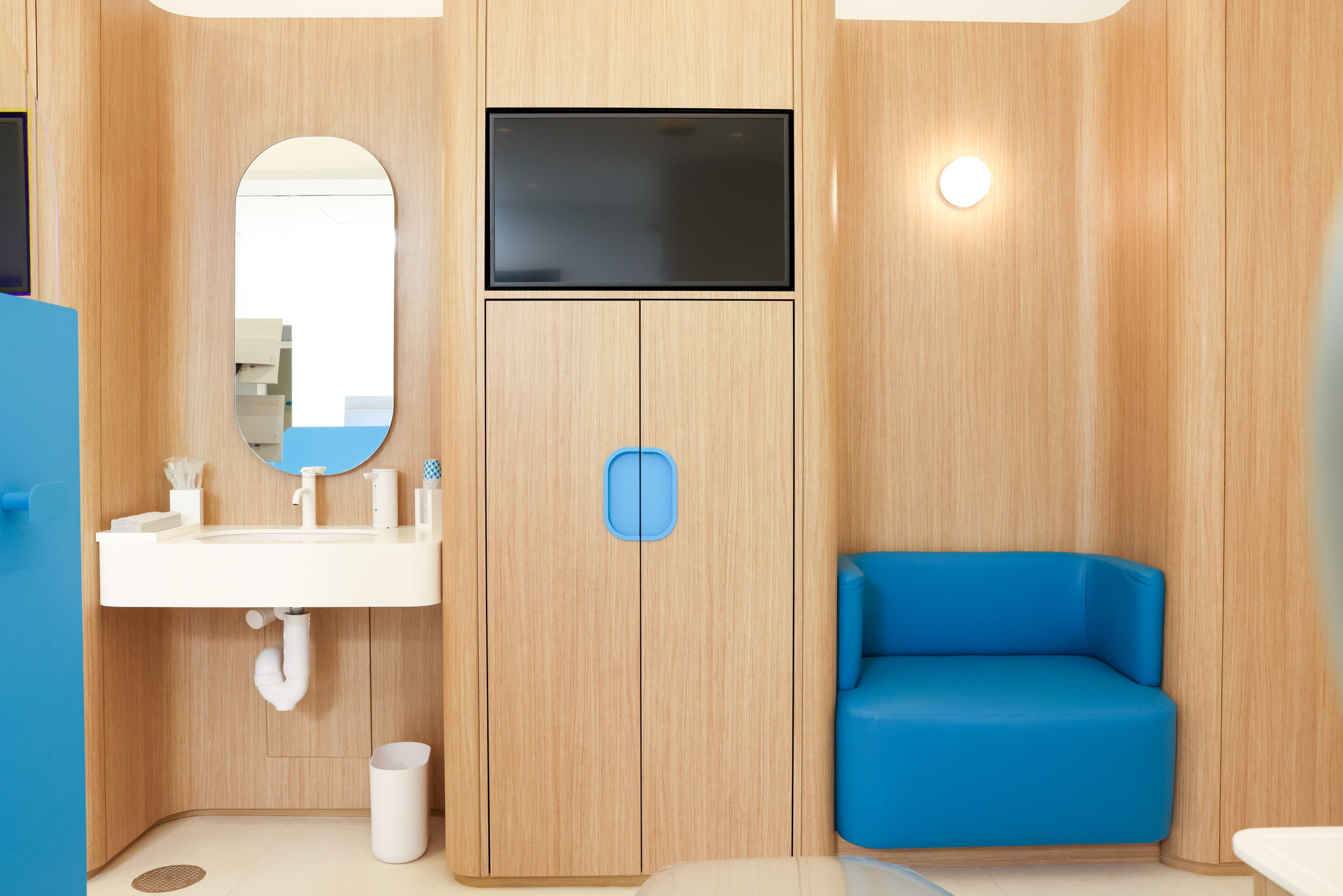 Contemporary dentist office interior with light wood wall panels and integrated cabinetry, featuring a mounted screen and oval mirror above a minimalist white sink. The design emphasizes clean lines and ergonomic layout, complemented by a bright blue chair for patient comfort. Soft lighting and natural wood tones create a calming, modern aesthetic that blends functionality with inviting architecture.