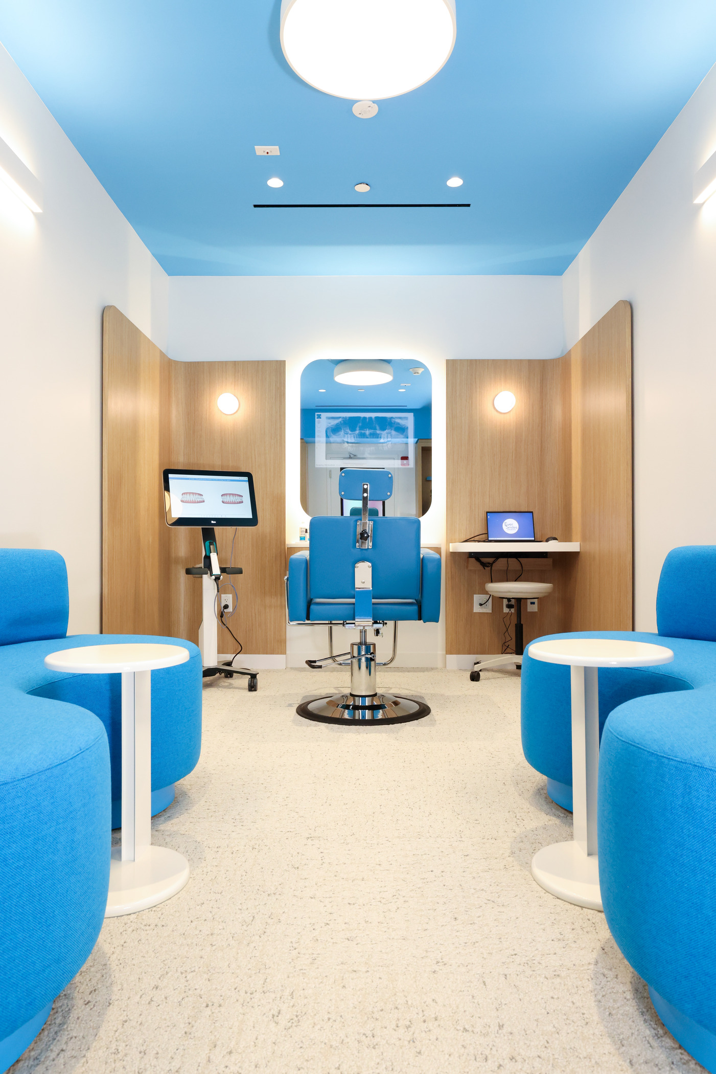Modern dentist office interior with a bright blue ceiling and matching seating, creating a vibrant and calming atmosphere. The space features ergonomic design with a central dental chair, flanked by wood-paneled partitions housing a computer workstation and diagnostic screen. Clean white walls, round ceiling light, and minimalist furniture emphasize a contemporary, patient-friendly layout.