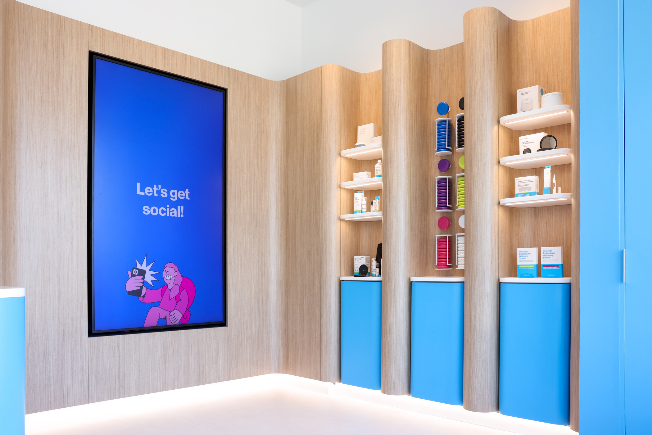 Dental office display area with light wood walls and bright blue cabinets. Shelves hold dental care products and colorful orthodontic samples. A large blue sign reads ‘Let’s get social!’ adding a modern, friendly touch.