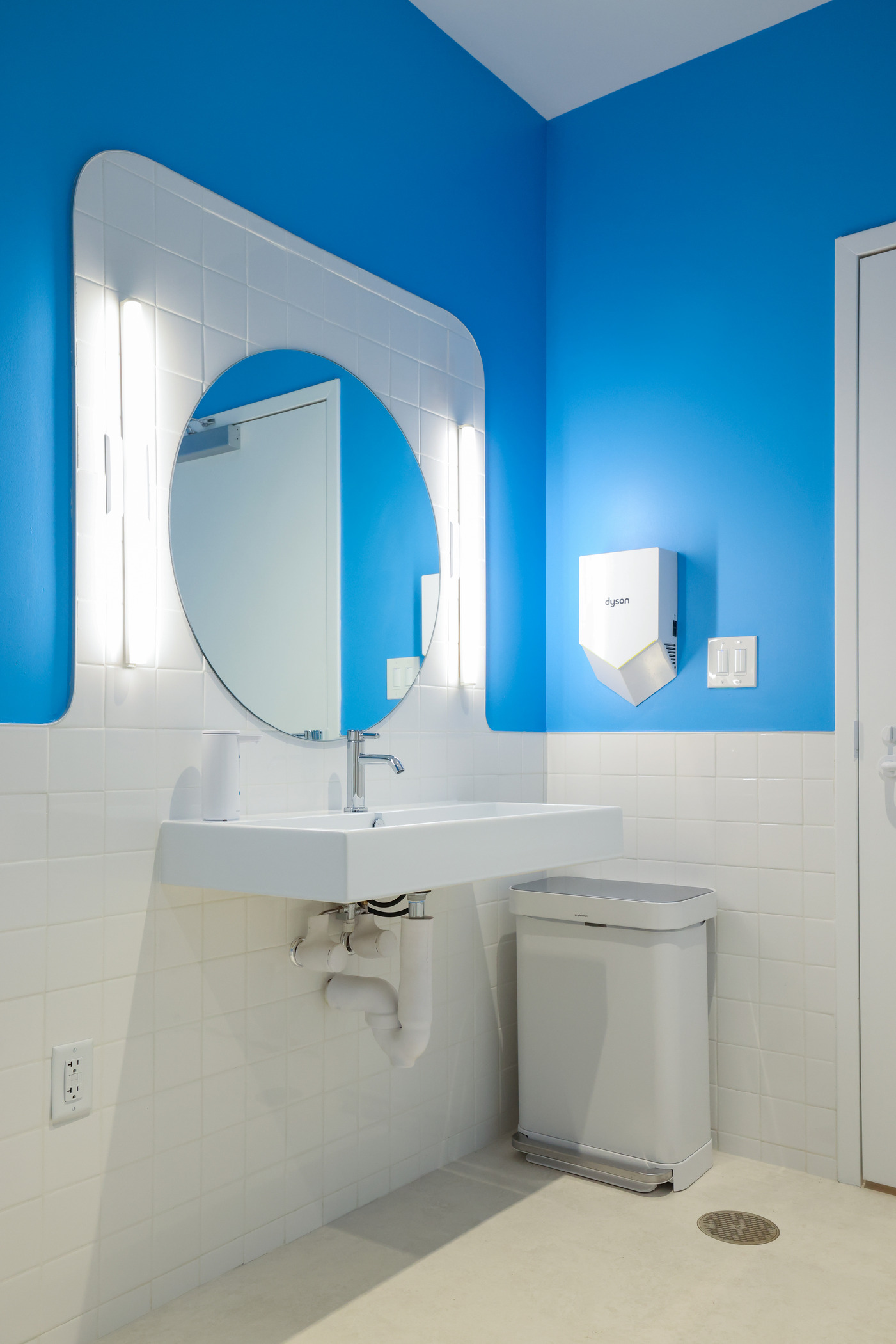 Dental office restroom with bright blue walls and white tile backsplash. Features a round mirror with built-in lights, a white sink, a trash bin, and a wall-mounted hand dryer for a clean, modern look.