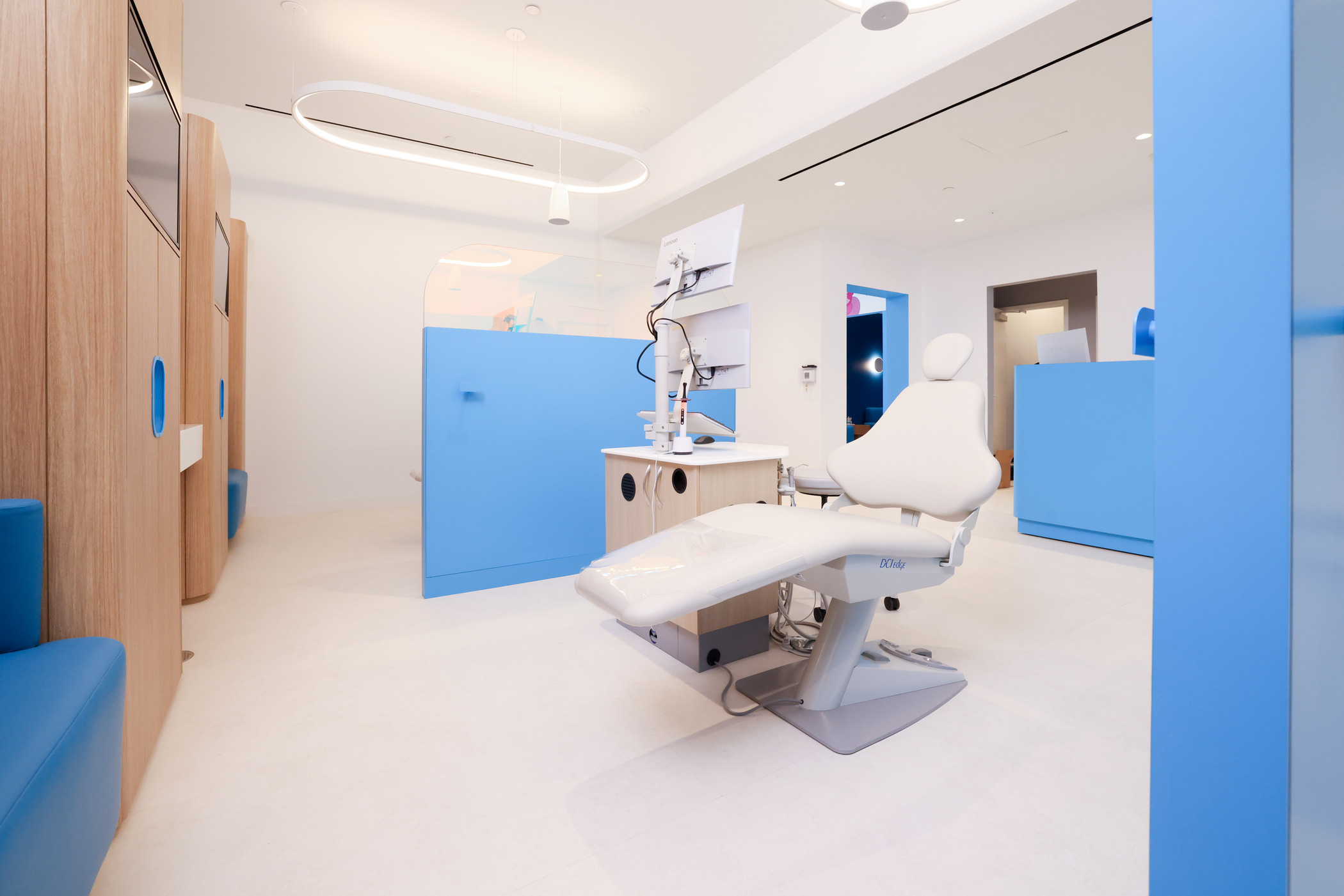 Dental treatment room with a modern reclining chair, bright blue partitions, and light wood accents. The space includes a mounted monitor and sleek overhead lighting, creating a clean and contemporary look.