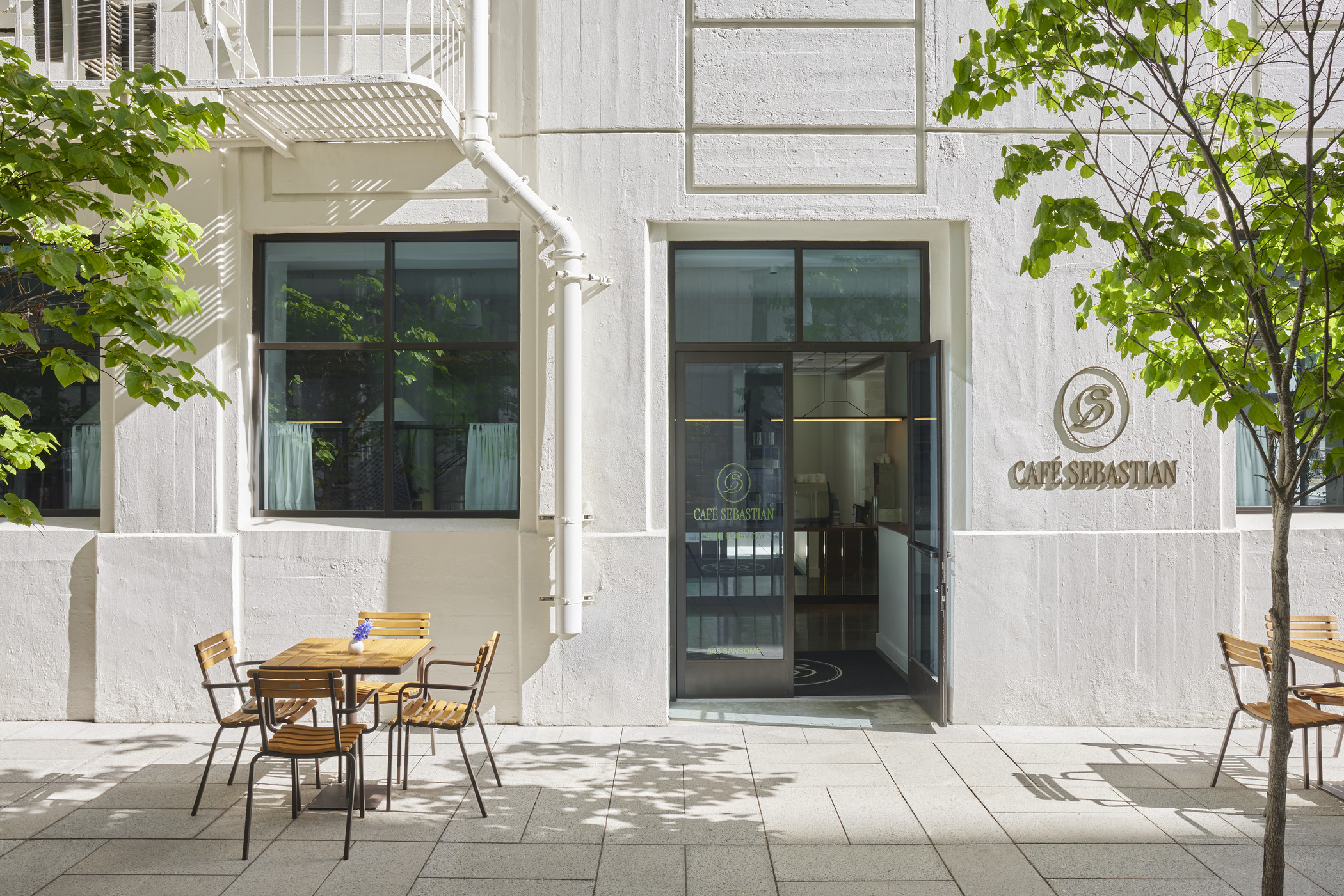 A cafe exterior is photographed in the daytime, with two sets of empty seating arranged around a stately stone entrance with young trees along the sidewalk