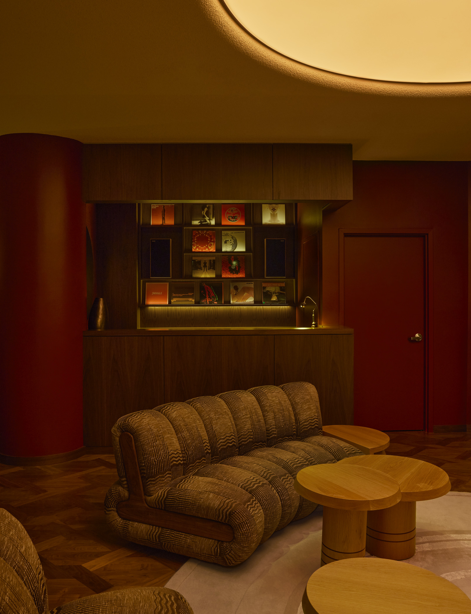 Modern lounge interior in a boutique restaurant with curved upholstered seating, round wooden tables with illuminated shelving displaying record albums. Elegant hospitality architecture combines warm tones and contemporary design elements.