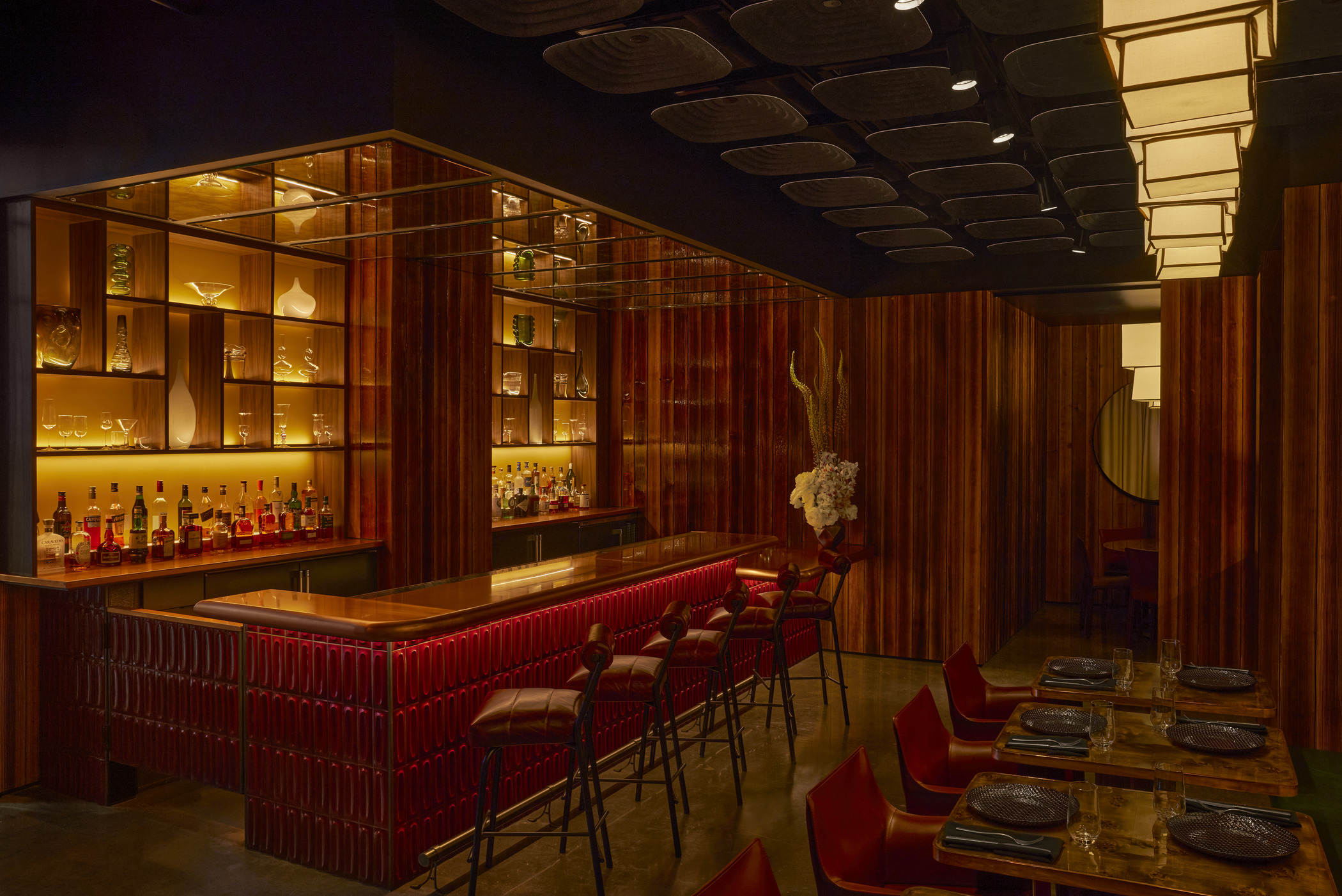 Restaurant bar with rich wood paneling, illuminated glass shelving, and red leather bar stools. Warm lighting and mirrored surfaces highlight contemporary restaurant architecture and upscale lounge design at Ama by Brad Kilgore.