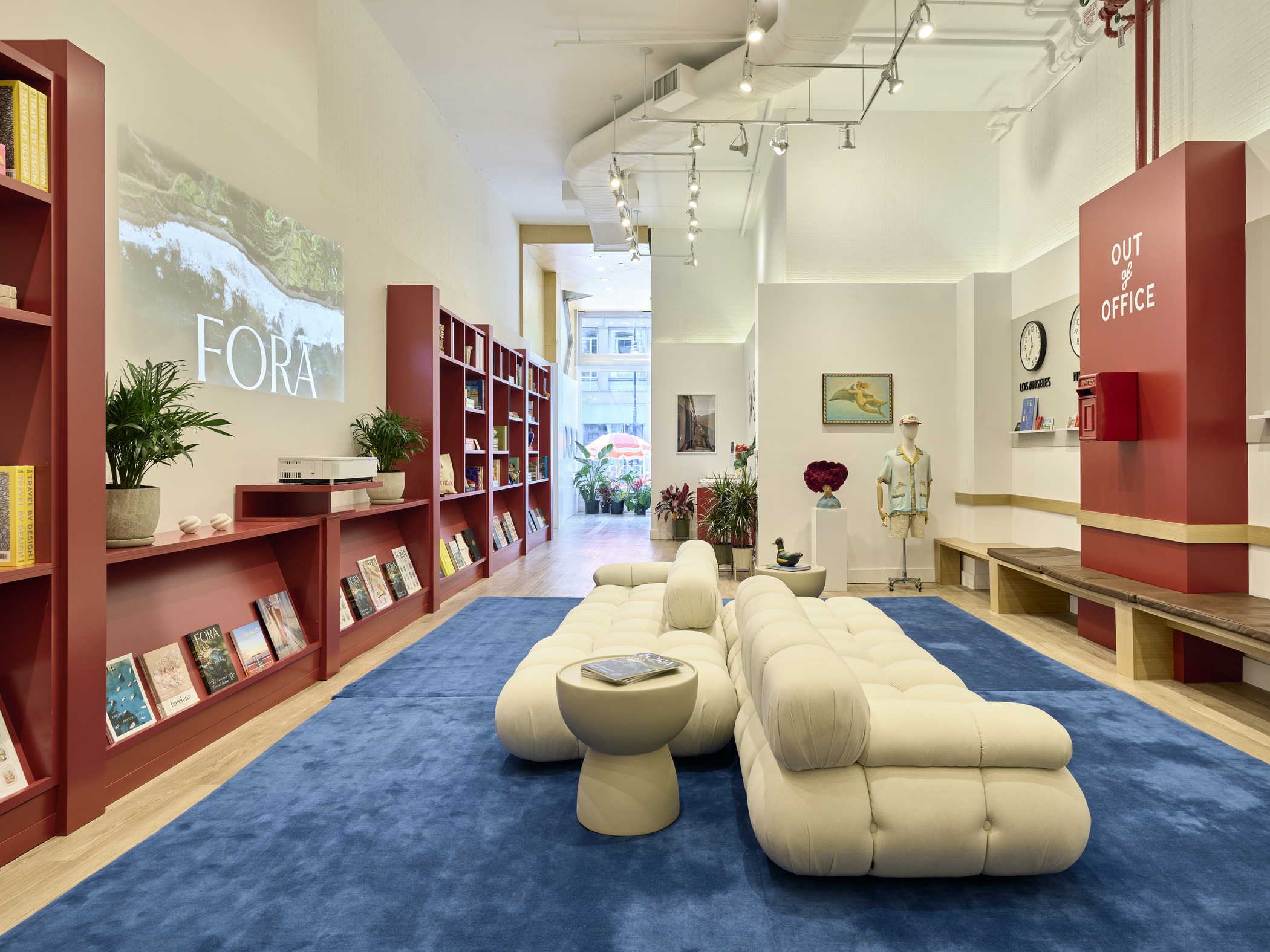 Fora Travel pop-up interior design in SoHo featuring signature red shelving with travel books and decor, cozy white modular sofa, and hospitality-inspired architecture by Lawrence Group.