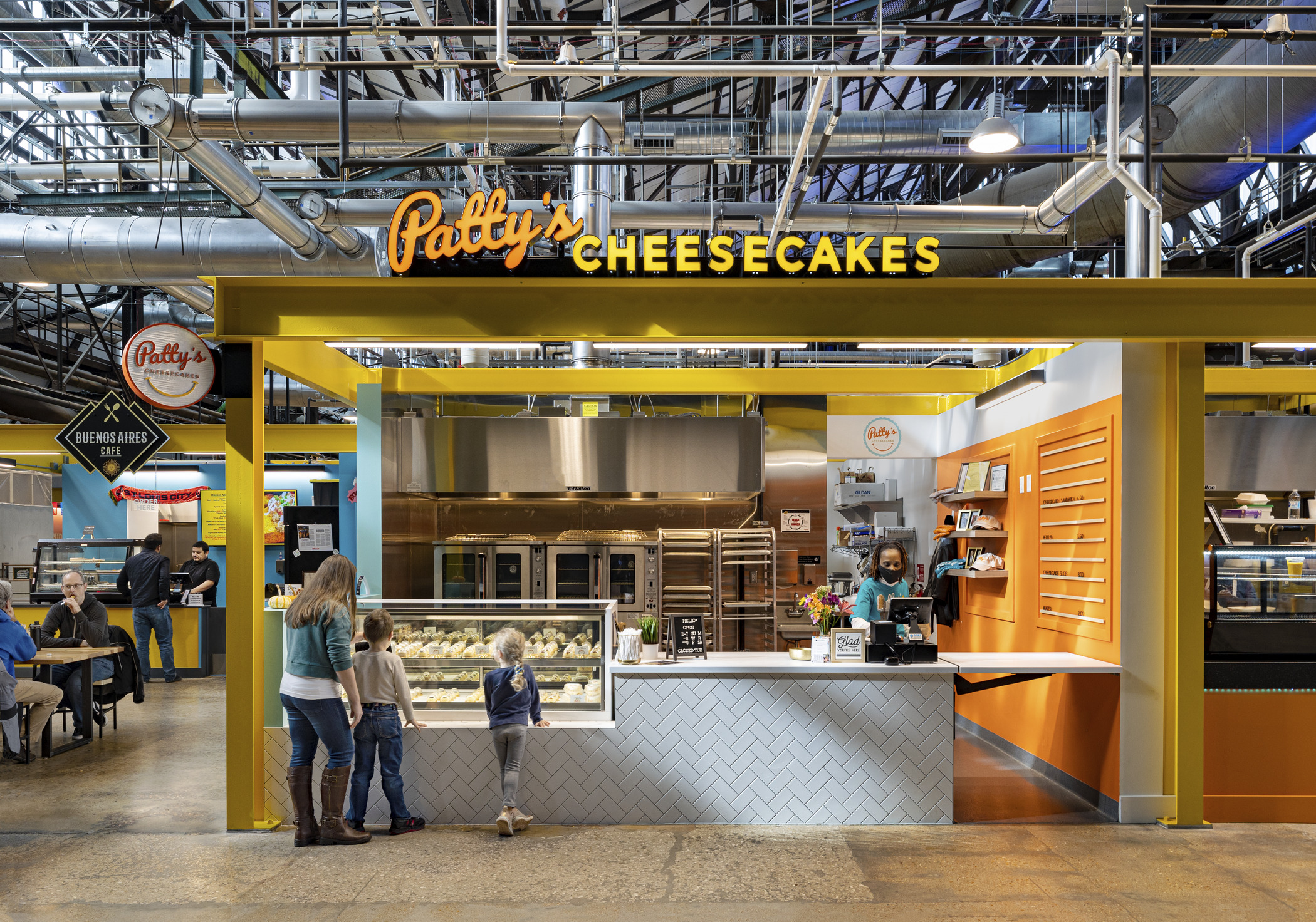 A mother and her two children order food at an open stall within a large former industrial space