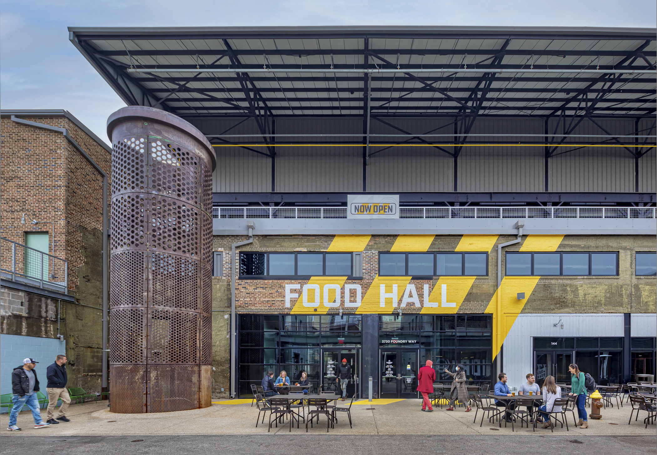 A variety of people are seated outside a large industrial building, with a color graphic saying "food hall" over the front entrance/wall.