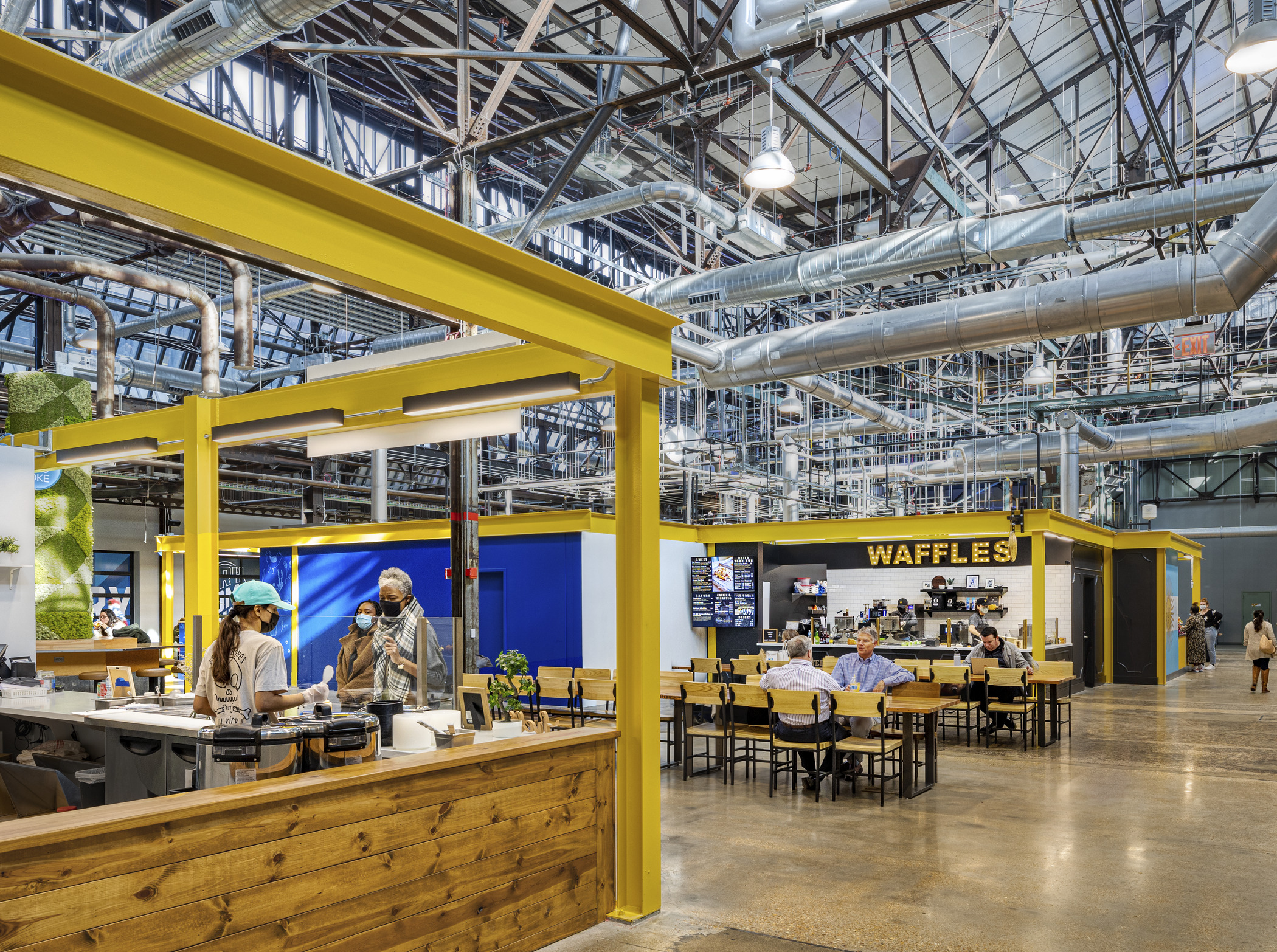 Two women order food at an open stall within a large former industrial space, as a variety of people eat at large picnic table and browse other food stalls