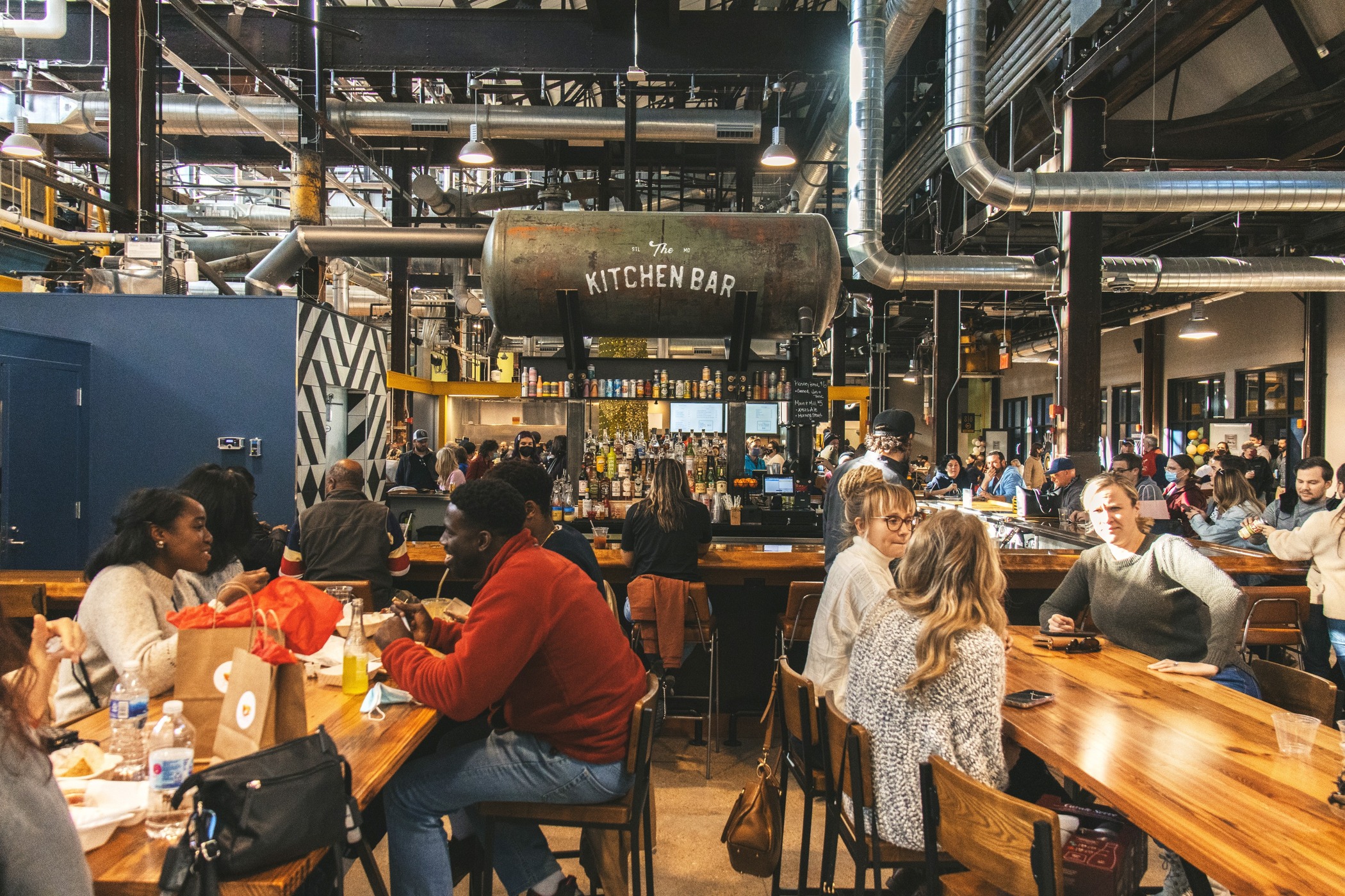 A variety of people eat at large picnic table-style bar and browse other food stalls within a large former industrial space
