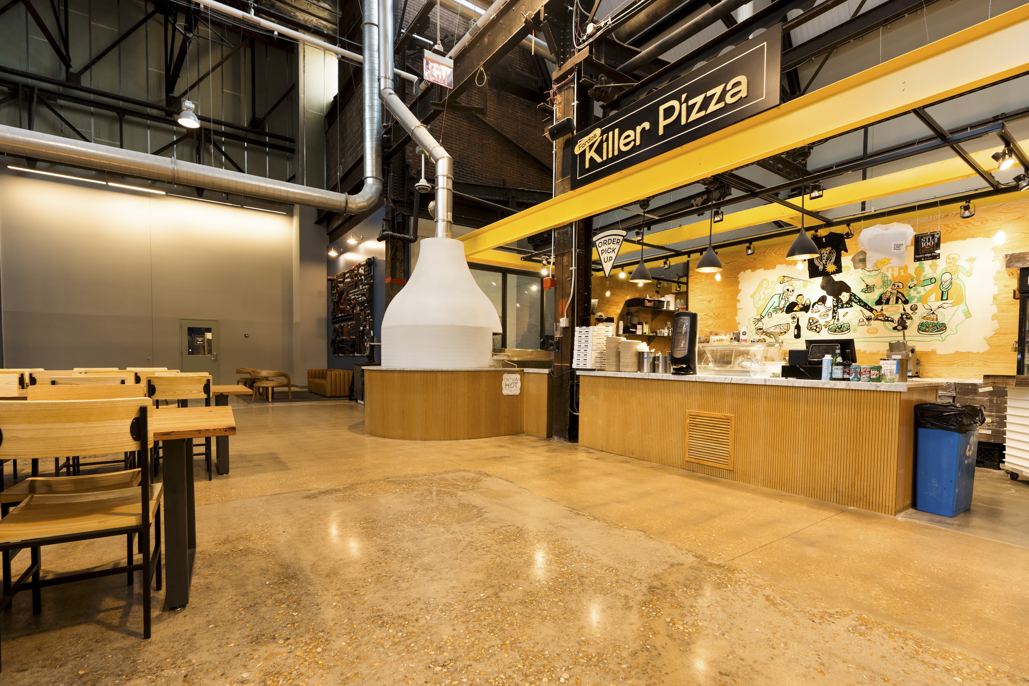 An open food stall and bright pizza oven are visible in a large former industrial space, which now has a variety of generous picnic tables to sit at