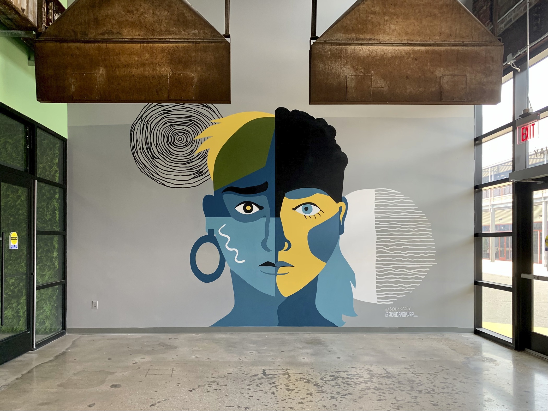Within a large, bright former industrial space, a select-tone mural of two androgynous faces are connected and split down the middle by a wavy line.