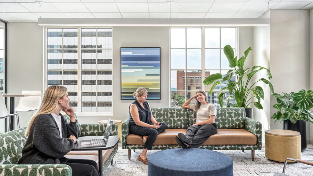 Three coworkers collaborating in a bright office lounge with biophilic patterned seating, large windows, and indoor plants.
