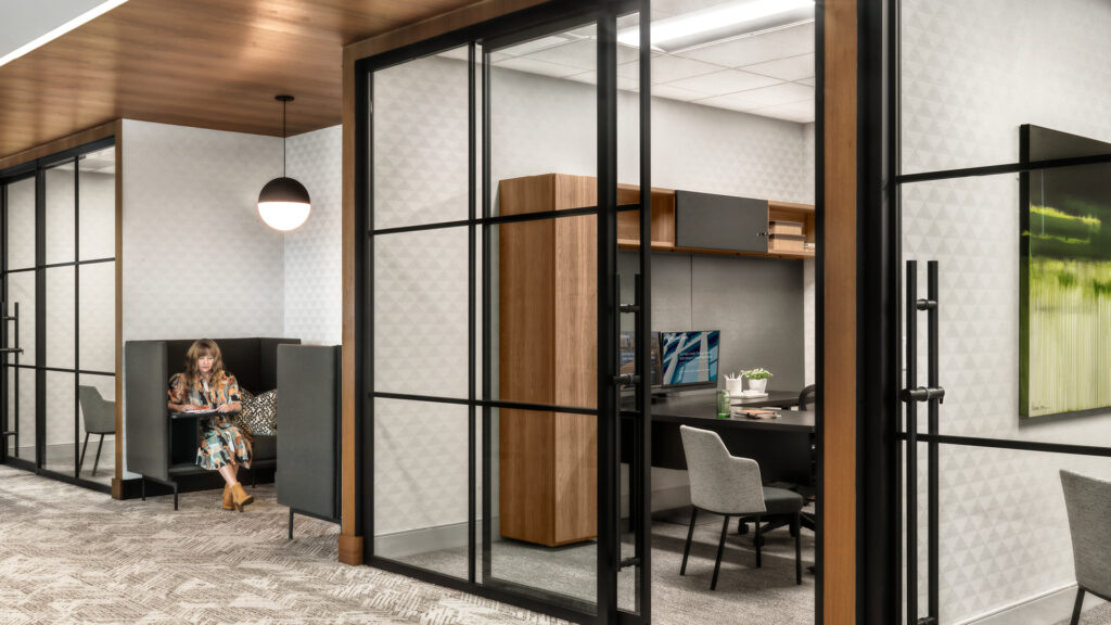 Employee working in an acoustic lounge booth beside glass-front private offices in a modern workplace corridor.